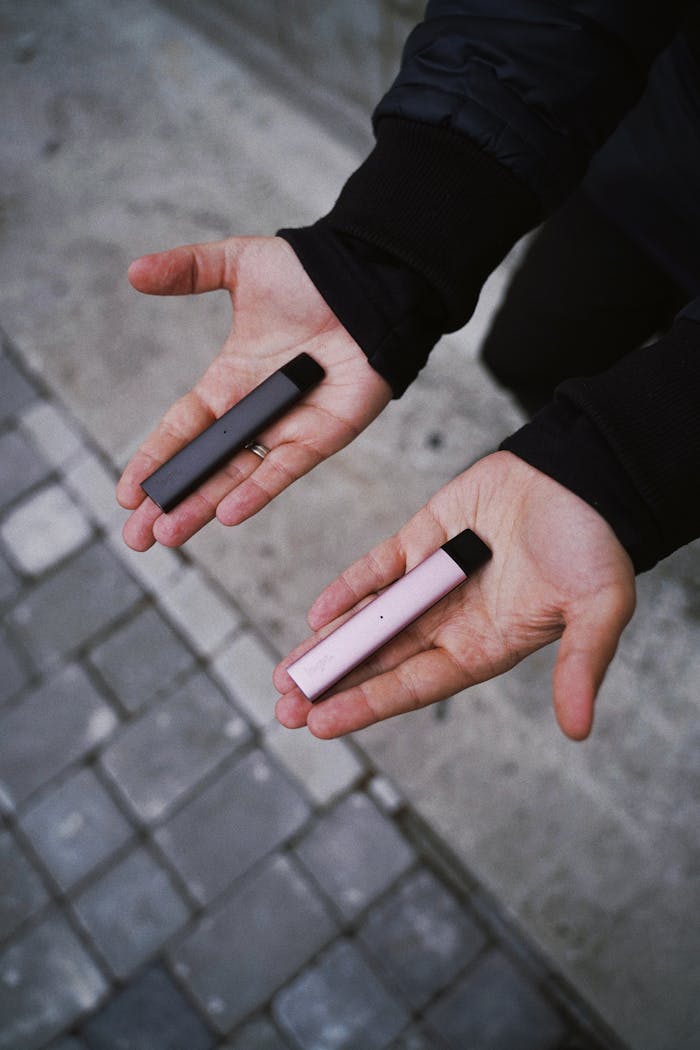 Close-up of hands holding two colorful vape pens on a city pavement.
