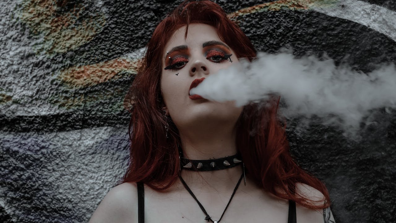 Portrait of a woman with red hair and bold eye makeup exhaling vapor against a graffiti wall.