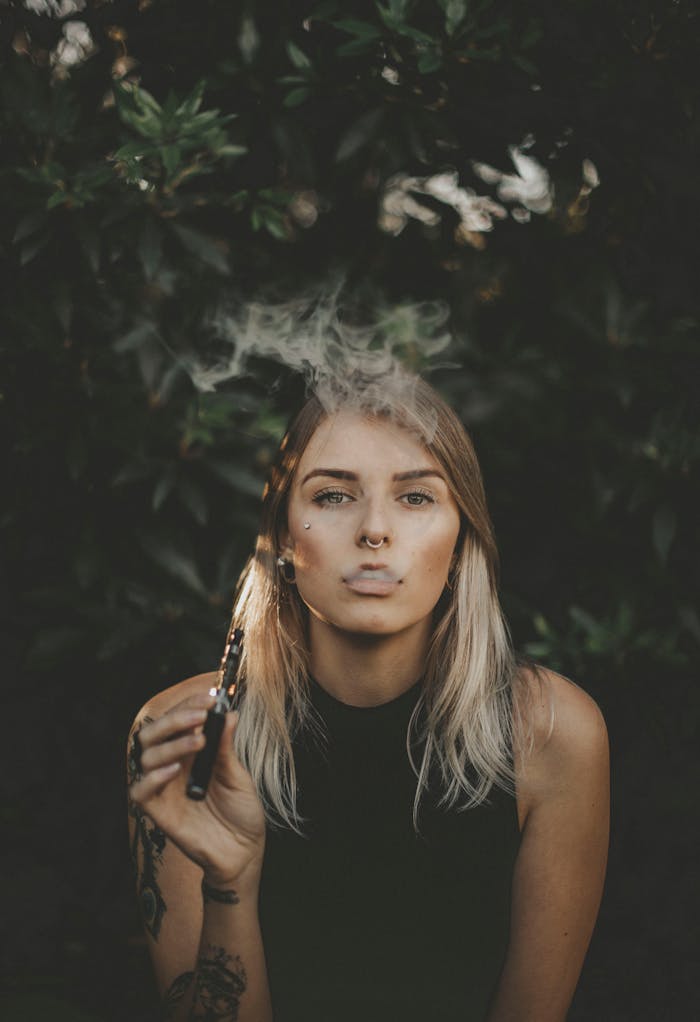 A young woman with blonde hair and nose ring is vaping outdoors, surrounded by greenery.