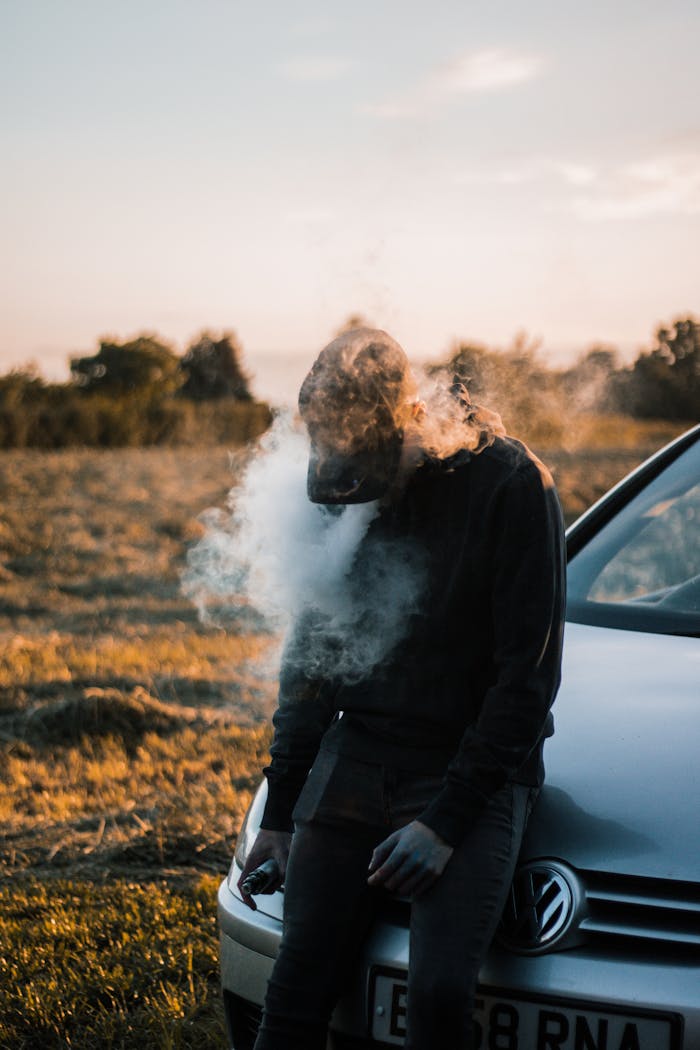 A man stands relaxed, vaping beside a car in a golden-hour field setting.