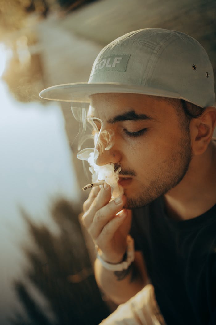 Contemplative young man smoking outdoors, captured in warm golden light, wearing a cap with visible nose piercing.